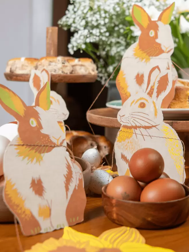 Three rabbits perched on table, with brown eggs and hot cross buns