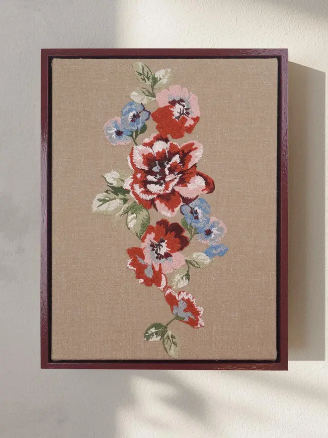 Framed embroidered floral artwork featuring red, pink and blue flowers on natural linen in a deep burgundy wooden frame.