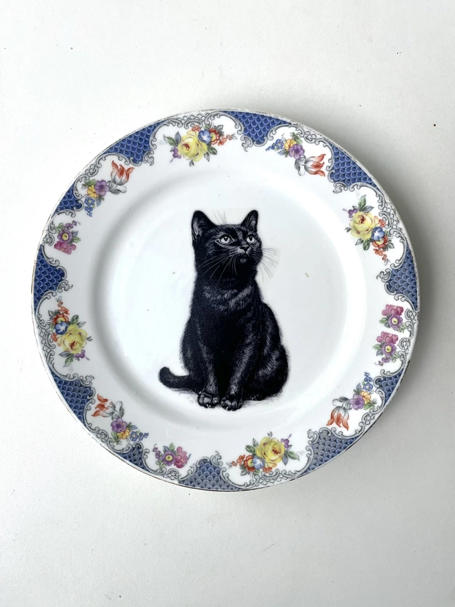 a vintage plate with an ornate border, in the centre of the plate is a vintage style black and white illustration of a black witch's cat