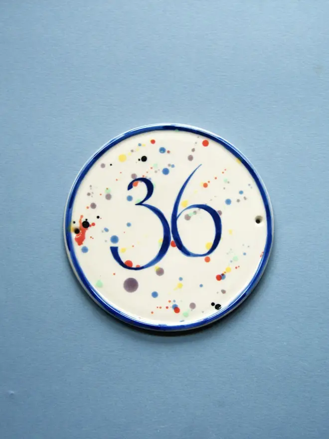 personalised hand-thrown ceramic house plaque. plaque features rainbow coloured dots and the number 36, photographed against a blue background.