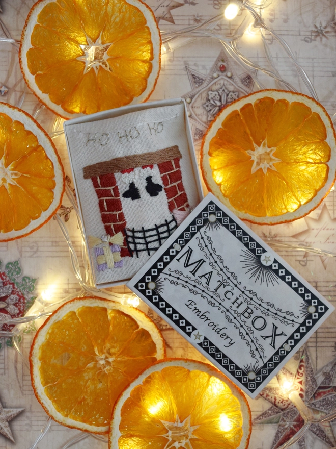 The fireplace embroidery, displayed in its matchbox.  Surrounded by dried orange slice decorations and fairy lights.