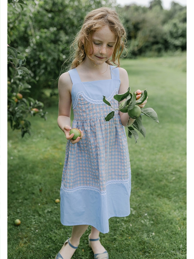 A girl holds an apple in a sundress with blue checked fabric, plain blue fabric and rikrak detail