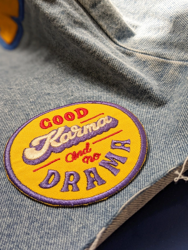 Yellow, Blue & Red round typographic patch reading 'Good Karma and No Drama'. The word Karma is written in a thick cursive lettering style in white, with a blue drop shadow, while the additional type is written in a retro style capital lettering & a smaller red cursive making up a fun retro style fabric patch. 