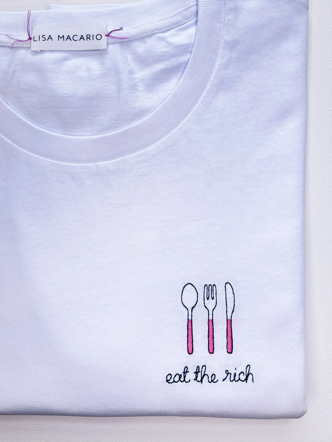     White T-shirt embroidered with pink cutlery motif and the words “Eat the Rich”