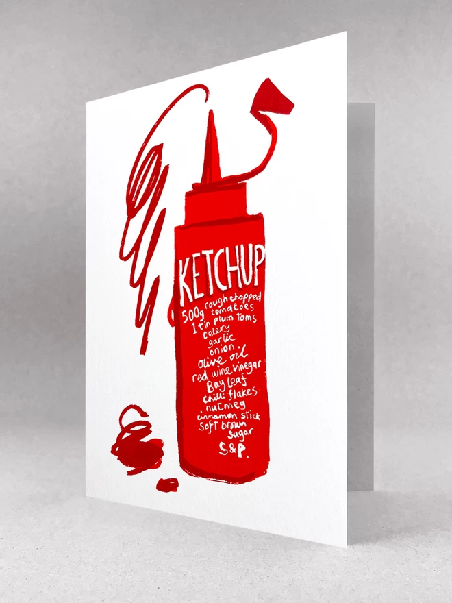 Ketchup recipe is written on a squeezy red bottle, with a scribbled squirt of ketchup jumping out the top - all on a white greetings card stood in a light grey studio space.