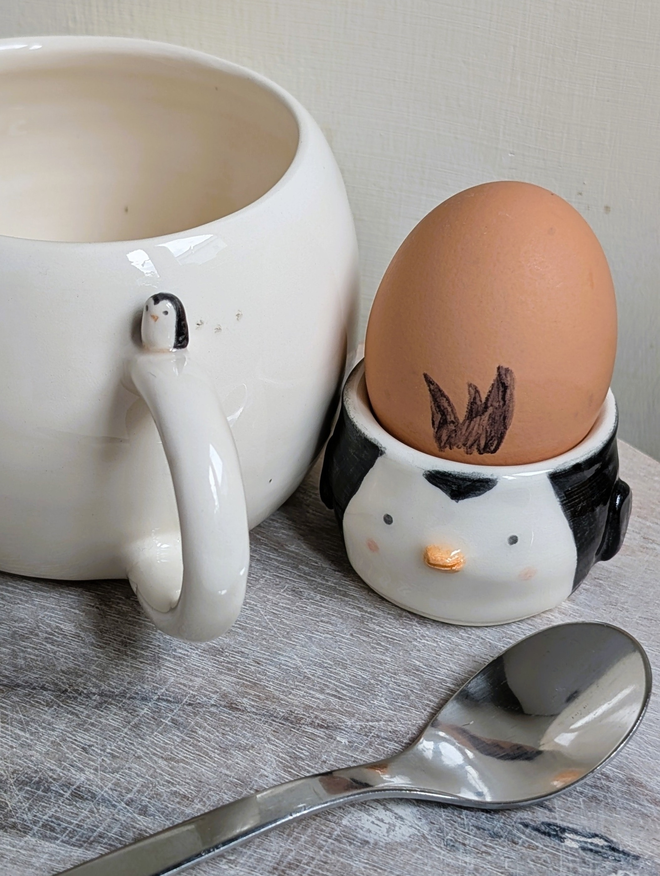 white cup with a tiny ceramic penguin on the handle next to a pottery penguin egg cup with an egg in