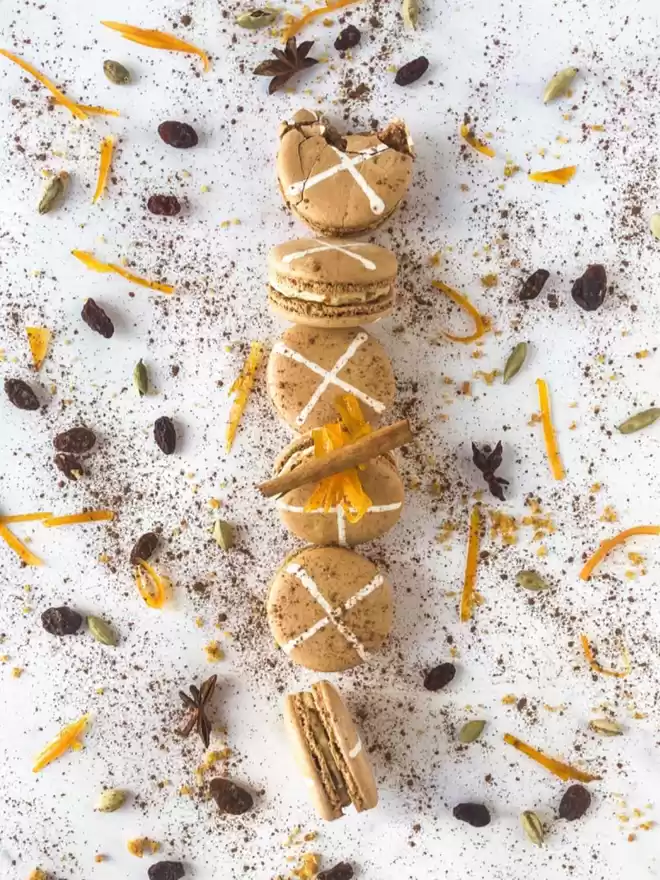 an assortment of hot cross bun macarons on a white surface with mixed spices and candied oranges