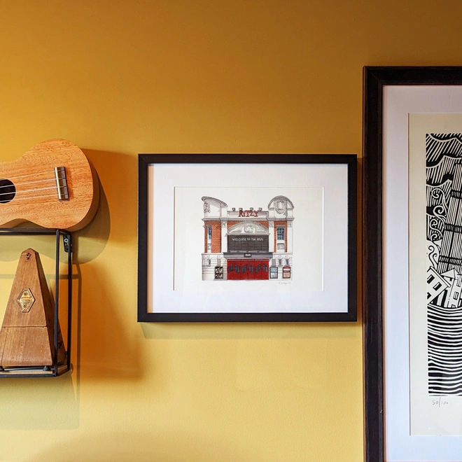 An illustration of the Ritzy Cinema in a black frame, hanging on a yellow wall beside a ukulele, a metronome, and another framed artwork.
