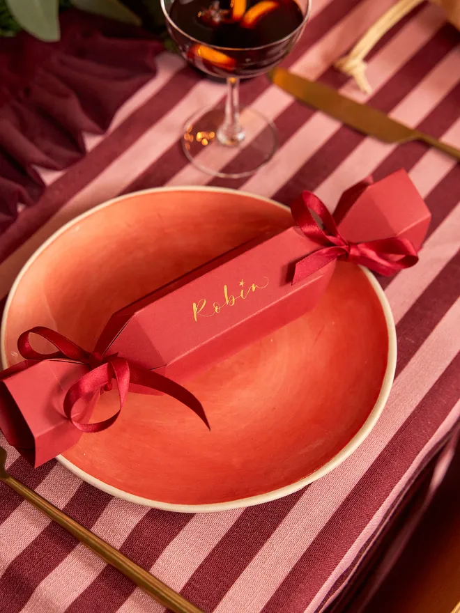 A personalised Christmas crackers in red with gold calligraphy with matching ribbon, atop a festive place setting on a red and light pink striped tablecloth