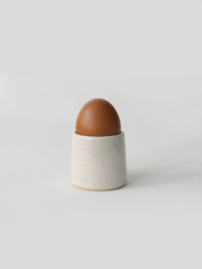 A handmade ceramic egg cup in speckled pepper white glaze holding a brown egg, photographed against a plain white background