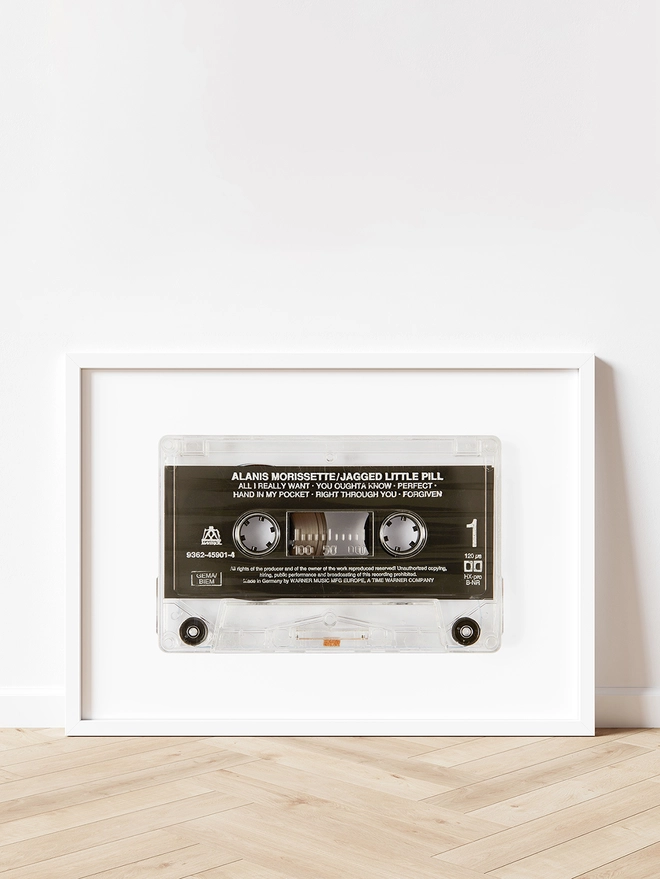 jagged little pill alanis morrisette cassette tape print. framed print propped up against white wall on wooden floor.