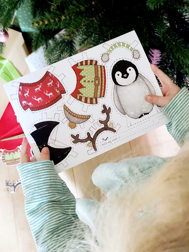 A child is holding a dress up penguin paper doll greetings card beside a Christmas Tree.