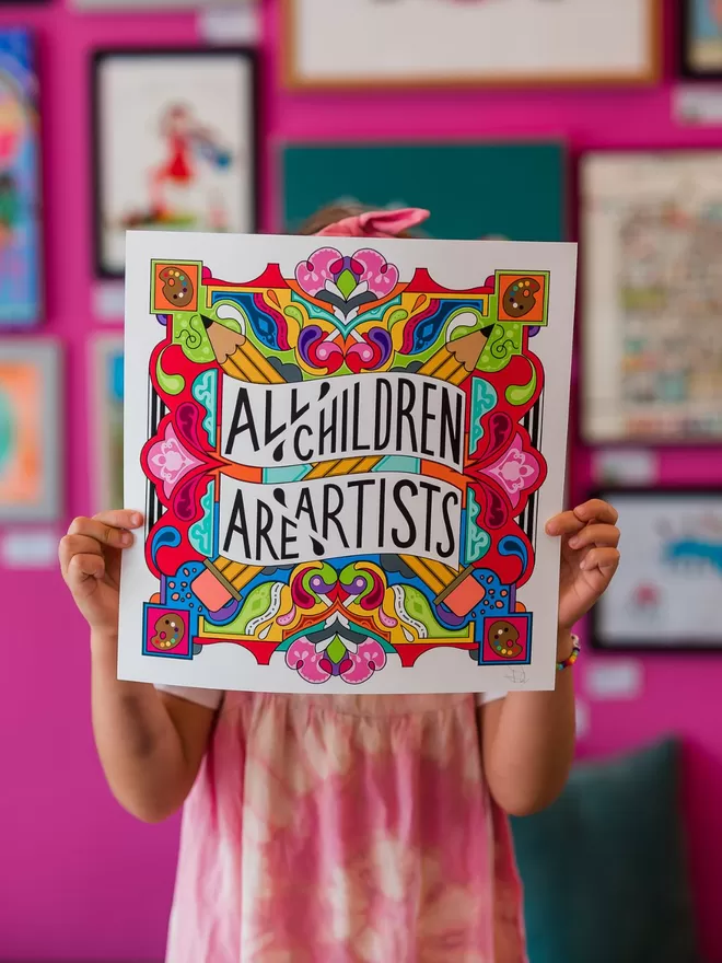 A child in a pink dress holding up a print saying 'All artists are children'