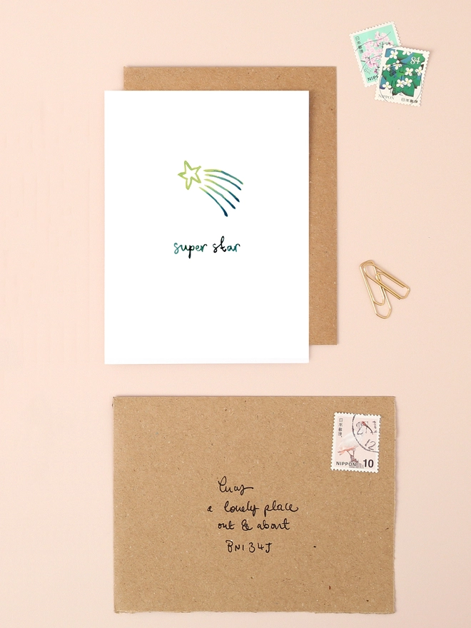 An image of a mini, rectangular white greetings card with an illustration of a shooting star in green ink. A hand written 'super star' message also in green ink is seen under the central illustration. The card is placed on top of a brown rectangular kraft envelope.