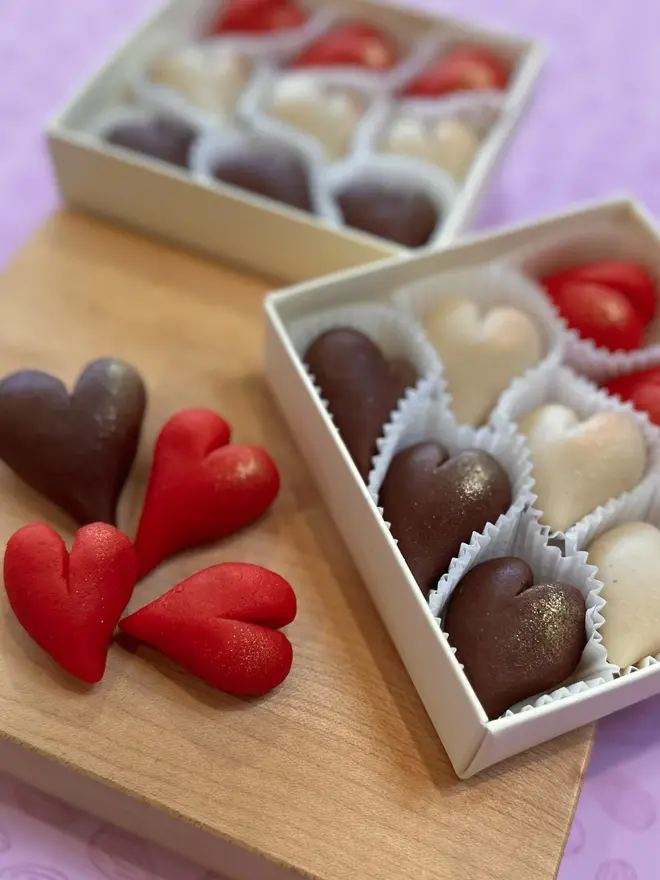 Marzihearts Valentines Chocolates (Box Of 6, 9, 12), two boxes of valentines chocolates on a wooden chopping board. The cherry, chocolate and marzipan chocolate hearts sit in front of a purple background. 