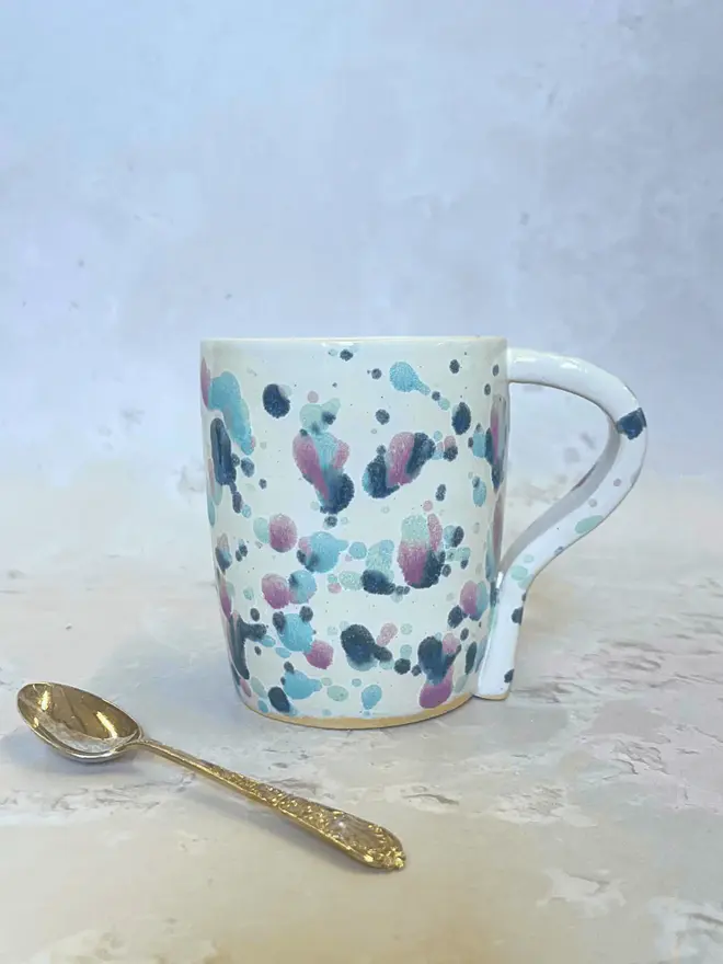 Sweetpea Standard Mug, a colourful mug placed on a stone surface in front of a plain backdrop. A metal spoon accompanies it. 