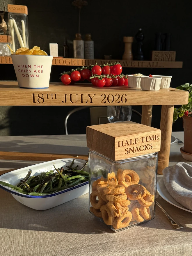 Personalised 'Half-Time Snacks' oak jar — a glass storage jar with an engraved oak lid, filled with savoury snacks and styled on a laid table beside a personalised oak serving bench.