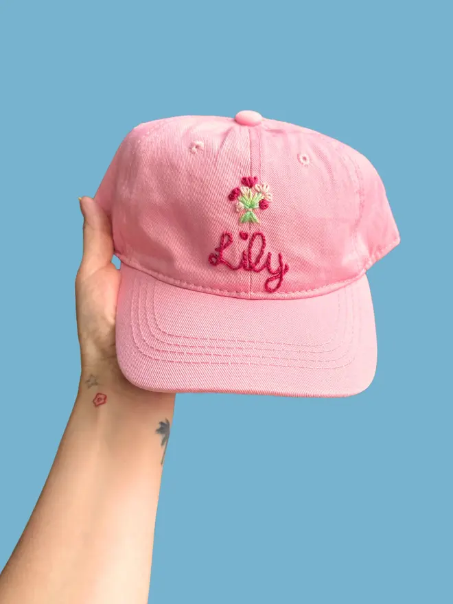 Hand Embroidered Kids Cap, a person holding a personalised cap against a colourful backdrop. It says a child's name. 