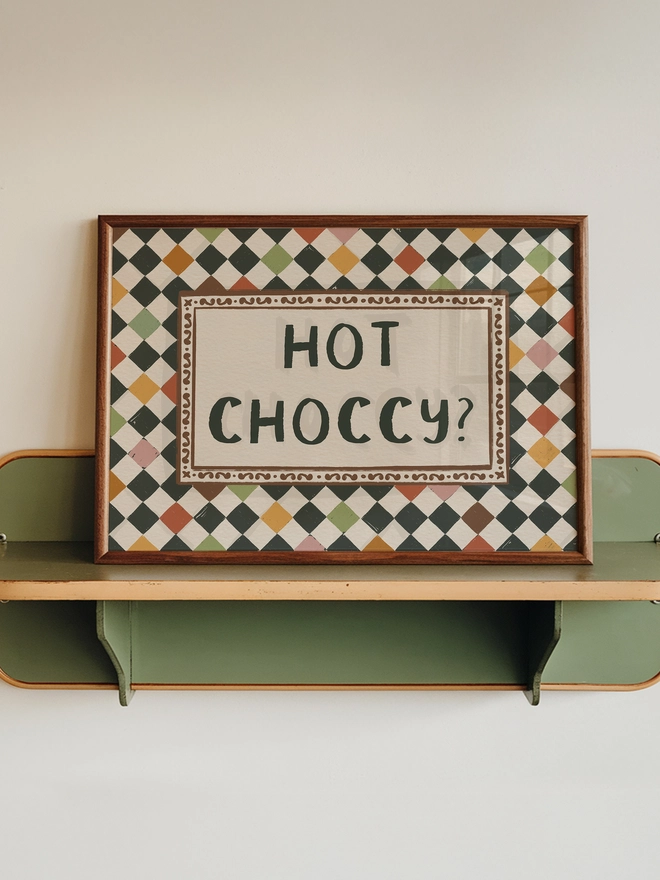 Checkerboard pattern with ' Hot Choccy?' text central, featuring a rich chocolate and jewel tone inspired colour palette, with a  harlequin check tile background. 