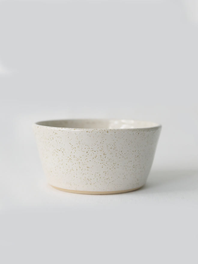 A single hand-thrown ceramic breakfast bowl in pepper white, with a glossy glaze showing subtle variation in tone, a gently flared form and unglazed natural clay base, photographed against a plain light grey background.