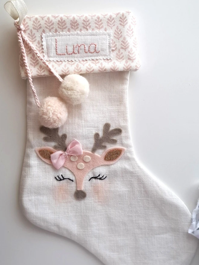 Traditional baby reindeer Christmas stocking in Traditional bay Christmas stocking in natural linen with a pink leaf pattern cuff, there are alpaca fluffy pompoms hanging at the side and a reindeer felt face.