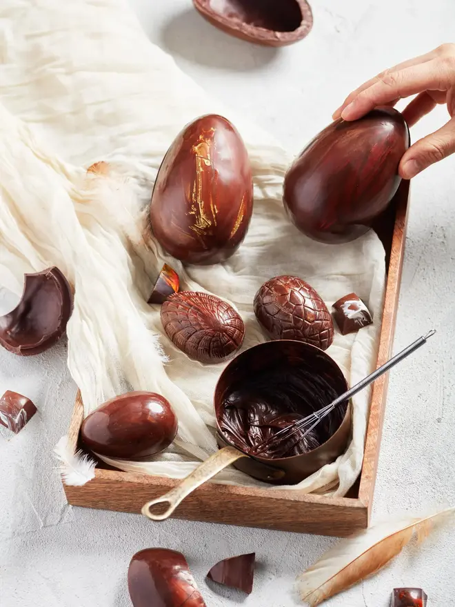 Extra Thick Milk Chocolate Easter Egg With Trevethan Gin, a person placing a chocolate egg on a wooden board. It is accompanied by an assortment of other chocolate eggs. 
