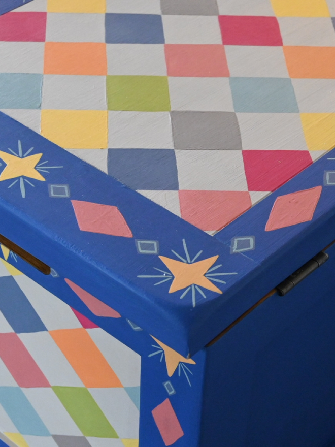 Close-up of the corner of a bright blue box or chest, showing a hinged lid with a repeating pattern of pastel-colored diamonds and a decorative border of smaller diamonds and yellow stars.