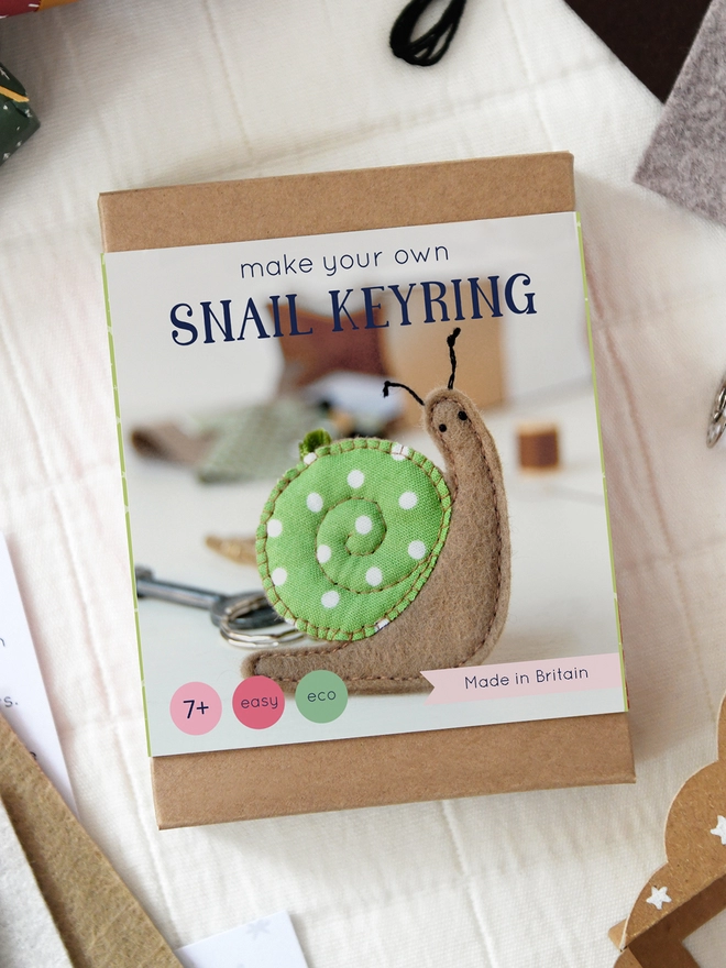 A snail keyring craft kit box lays on a fabric surface with various items from the kit laying beside it.