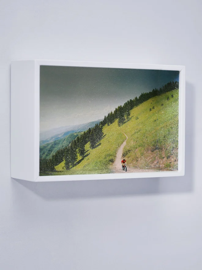 Miniature scene in an artbox showing a tiny man biking against a sparkling forested mountain backdrop.