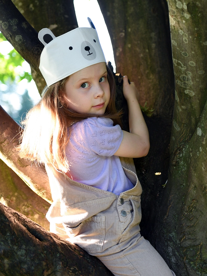 A young girl wearing a dress-up polar bear fabric crown and beige dungarees is climbing a tree.