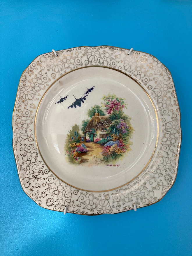 A gold and white china plate featuring a cottage with fighter jets in the sky.