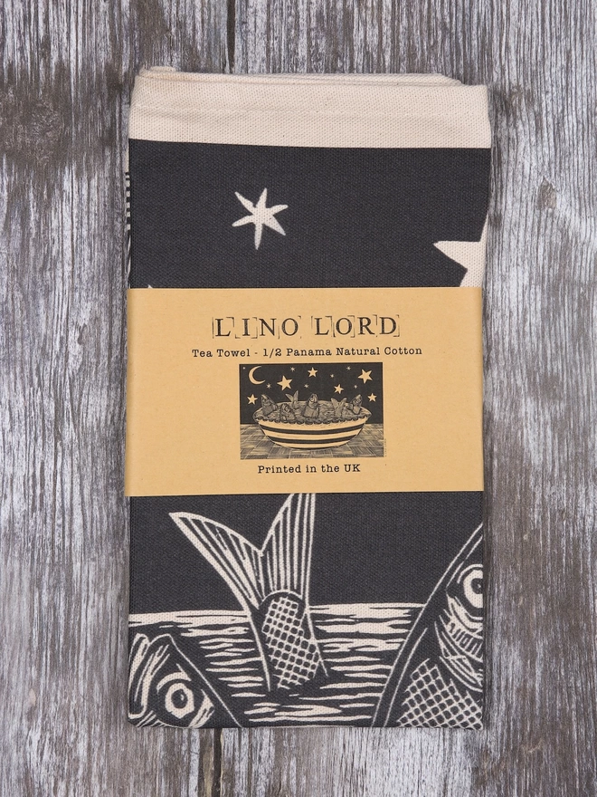 Picture of a folded tea towel with an image of a fish pie with the fishes gazing up at the stars, taken from an original lino print