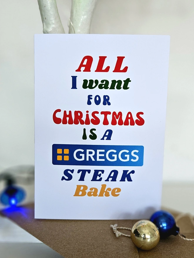 A colourful card Christmas Card that says All I want for Christmas is a Greggs Steak Bake in retro style fonts on a white background.