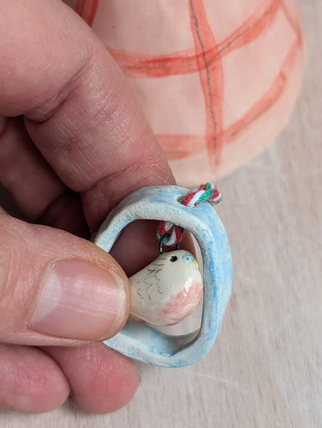 close up of a pink ceramic budgie that hangs from a pink and red check pottery bell