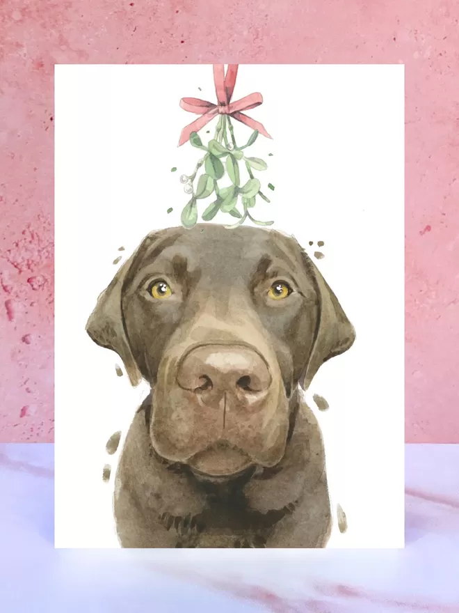 A Christmas card featuring a hand painted design of a Chocolate Labrador, stood upright on a marble surface.
