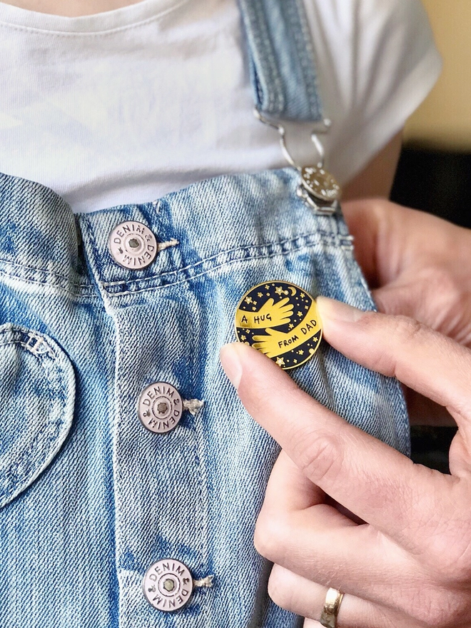 A Dad is pinning a navy and gold hug from Dad pin badge onto child wearing denim dungarees 