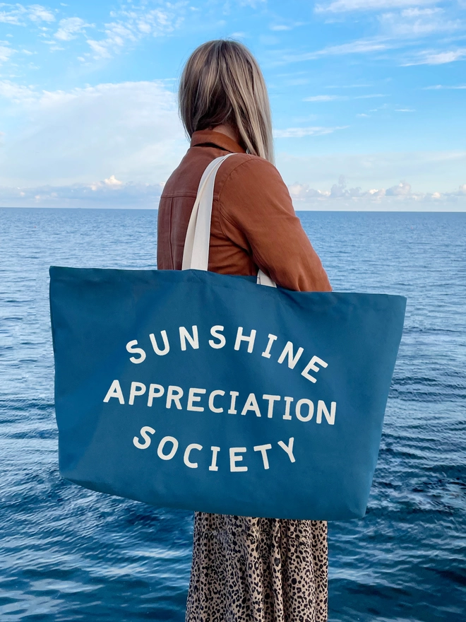 Model holding an blue oversized tote bag with Sunshine Appreciation Society slogan in front of the sea