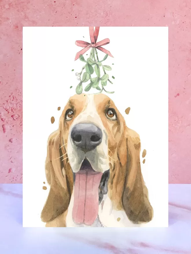 A Christmas card featuring a hand painted design of a Basset Hound, stood upright on a marble surface.