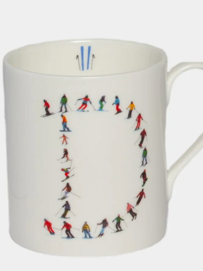 A simple white mug illustrated with a ‘D’ made of small, multicoloured skiers. The mug is set against a plain, white background.