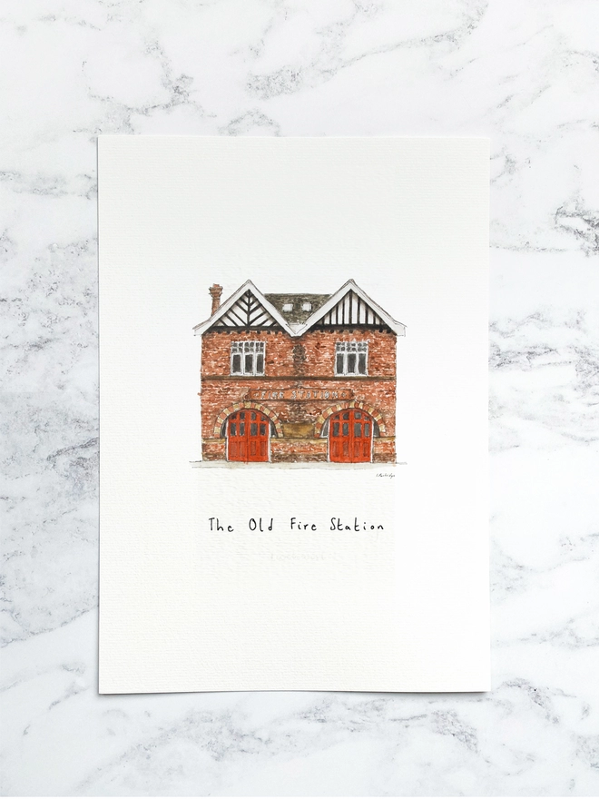 Beautiful watercolour illustration of The Old Fire Station in Tonbridge.  A brick building with two red framed arched double doors. The watercolour style is painted with a black pen outline and organic loose style with small details. The print is a small illustration on the centre of a white page and the paper sits on a white marble background.