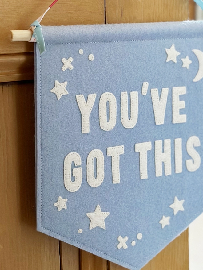A blue felt wall hanging, with the words You’ve Got This and white felt stars, hangs from a ribbon hanger on a wooden door handle.