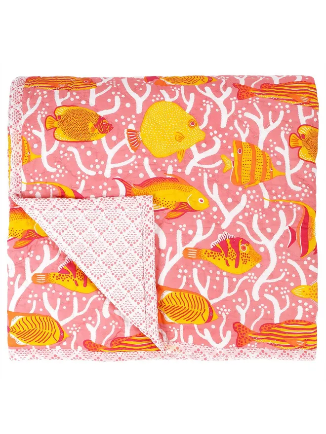 Pink Coral Reef Quilt, a pink quilt with a yellow and orange tropical fish print, folded on a white surface. 