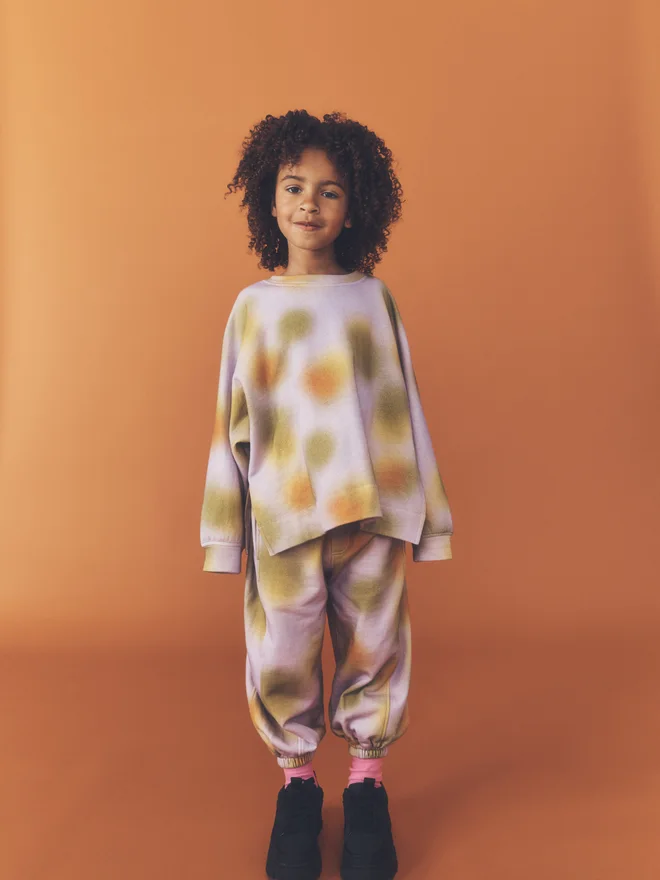 Child stands facing camera against a burnt orange backdrop, wearing the Studio Koter Riverson top in Hazy Lilac with matching print joggers.