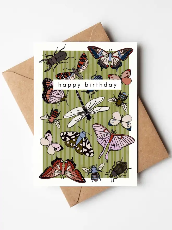 A colourful illustrated birthday card featuring butterflies, bees, beetles, dragonflies and other insects on a soft green striped background, with the words "happy birthday" across the centre. The card is displayed with a kraft envelope.