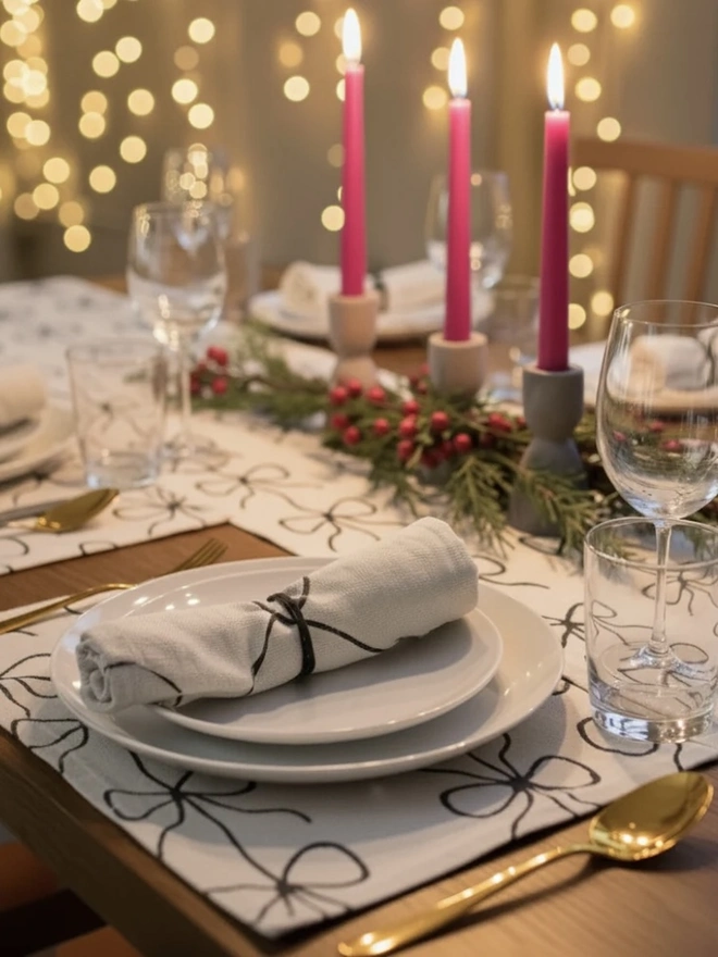 Hand Painted Bow Print Sustainable Linen Table Runner, a table set with plates and candles accompanied by a table runner. 