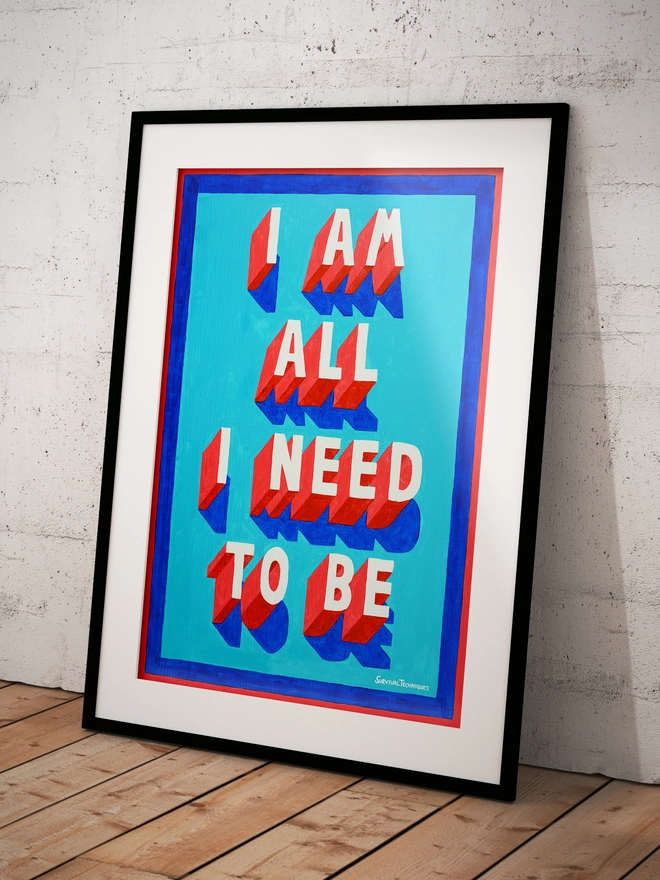 Framed typographic art print leaning against a white textured wall on a wooden floor. The artwork features bold 3D-style lettering in red with white faces and blue shadows on a bright turquoise background. The text reads "I AM ALL I NEED TO BE" in uppercase letters, framed by a red and blue border. Signed "Survival Techniques" in the bottom right corner.