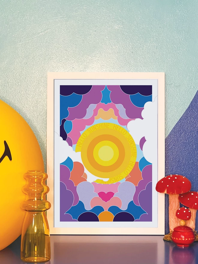 A concentric yellow circle sun with Hello Sunshine repeated three times in the outer ring sits at the centre of this portrait illustration. Surrounding it is an abstract design of clouds in purples, greys and oranges. The picture is in a white frame leaning against a painted blue wall on a blue cabinet. Next to the frame is a large illuminated yellow Smiley lamp, a yellow vase, a model of three red and white toadstools and a small round red wooden pot.