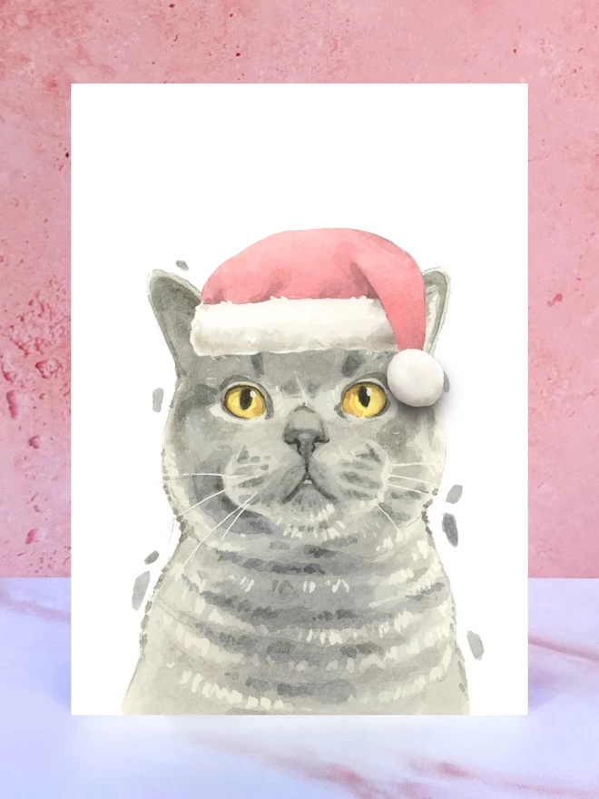 A Christmas card featuring a hand painted design of a british shorthair cat, stood upright on a marble surface surrounded by pompoms. 