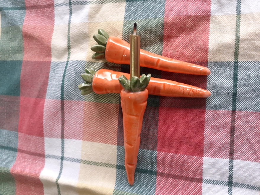 Three carrot-shaped ceramic candleholders arranged with a birthday candle inserted into one, displayed on a soft spring-toned fabric background.