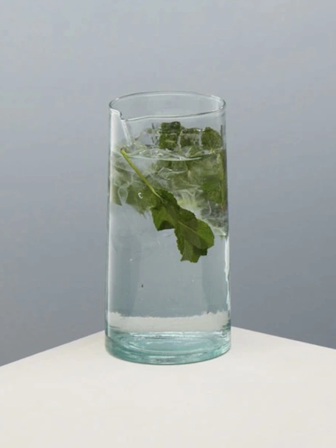 Glass Carafe, a large glass jug sitting on a plain counter top against a baby blue backdrop. It is filled by a soft drink. 